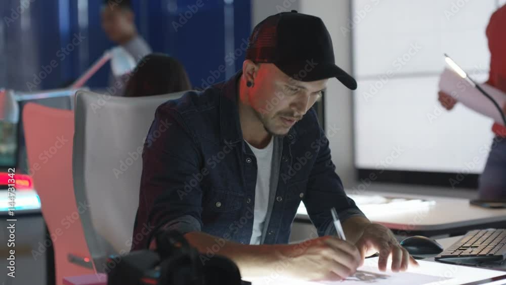  Young computer game designer drawing at his desk with colleagues working in background. 