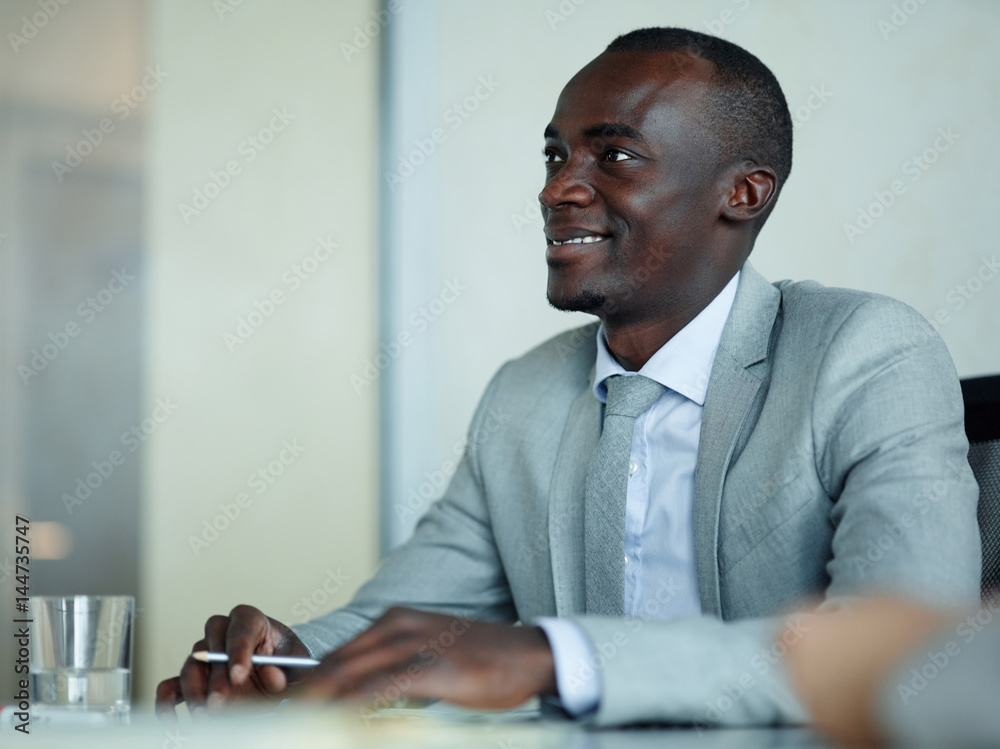 Portrait of successful African-American businessman sitting at meeting table in modern office, smiling and listening to colleagues intently Portrait of successful African-American businessman sitting at meeting table in modern office, smiling and listening to colleagues intently