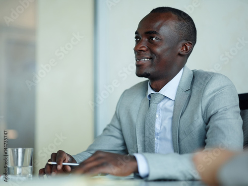 Portrait of successful African-American businessman sitting at meeting table in modern office  smiling and listening to colleagues intently