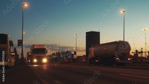 Semi trailer trucks and tanker trucks traffic passing by on street at dusk. Logistics transportation, container terminal in the background