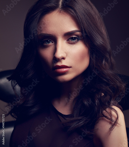Young cute makeup model in brown dress and curly hairstyle posing on dark deep shadow background. Toned closeup dramatic portrait