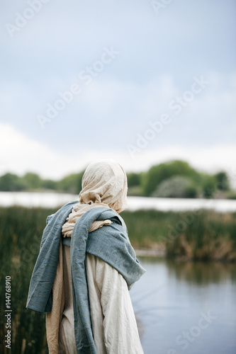 Jesus Christ pondering while standing next to a river, overlooking the setting sun. 