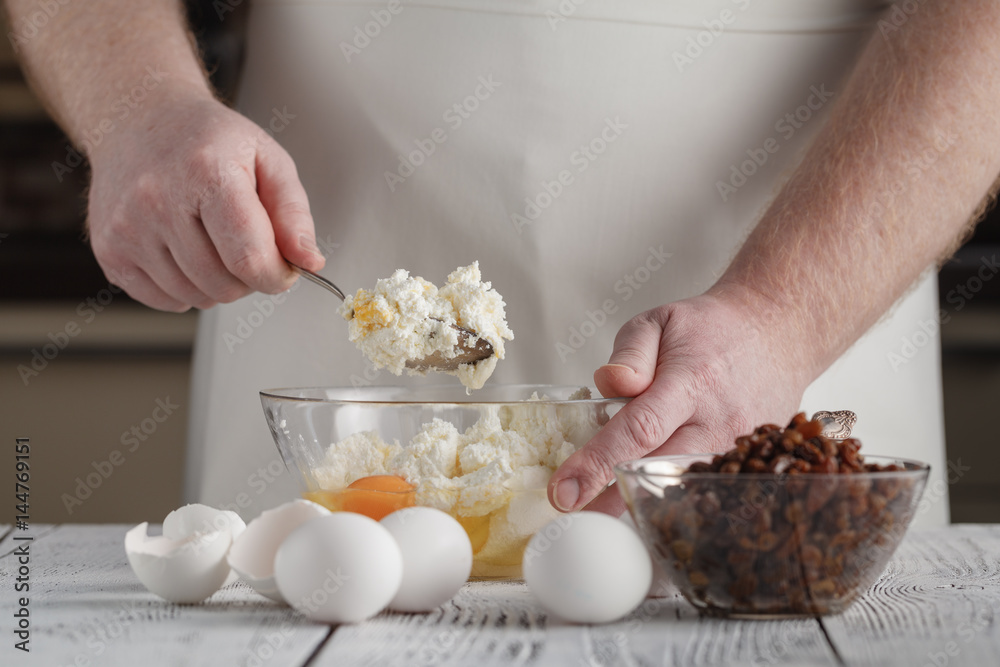 man's hands mixing cottage cheese, eggs, flour, raisins in the bowl on the table in the kitchen. Tasty, sweet meal. Healthy eating and lifestyle.