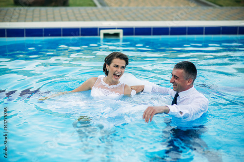 Young couple jumping in the swimming pool in a wedding suit and wedding dress. Sunny day. © Aleksandr