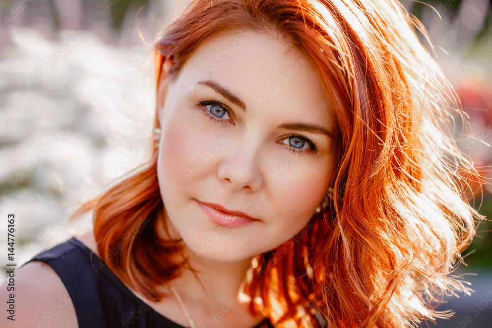 Fototapeta premium Closeup portrait of middle aged white caucasian woman with waved curly red hair with blue eyes, in black dress, looking in camera, outside in park, beauty fashion lifestyle concept