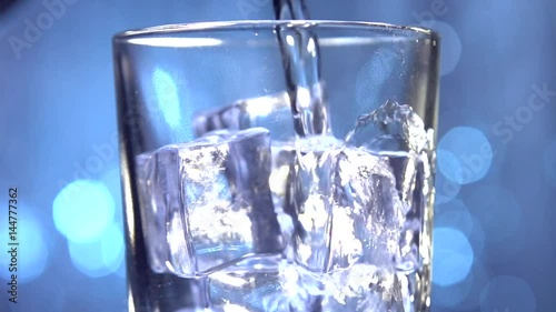 Water is pouring into a glass with ice cubes. Clear water pours ice cubes on a blue flickering background. Pure water  with Ice and bubbles in glass. Full HD  footage 1920x1080p. Slow motion 240 fps