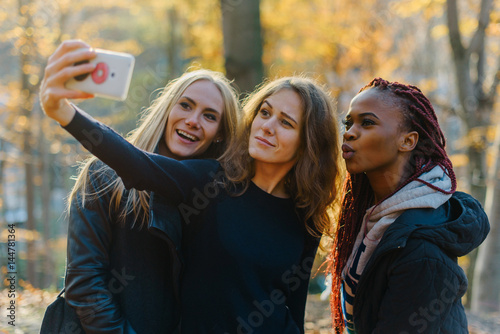 Three attractive happy woman making selfie in autumn park. Pretty girls with different colored skin. Female making funny faces photo