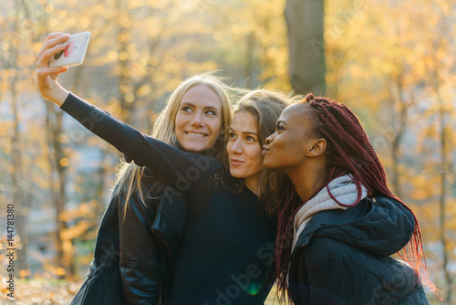 Three Pretty female making selfie in autumn park. Cute girls with different colored skin. Female making funny faces and smiling at camera. Sunny autamn day photo