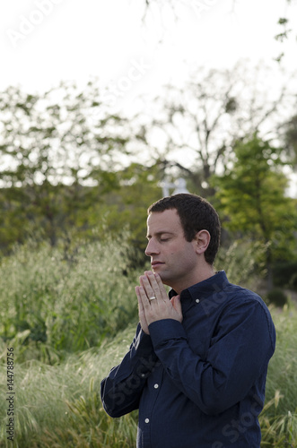 Praying man outdoors in a grassy field with hands together wearing a navy blue shirt.