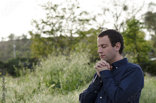 Married man praying outdoors in a grassy field with hands clasped up to his chin.