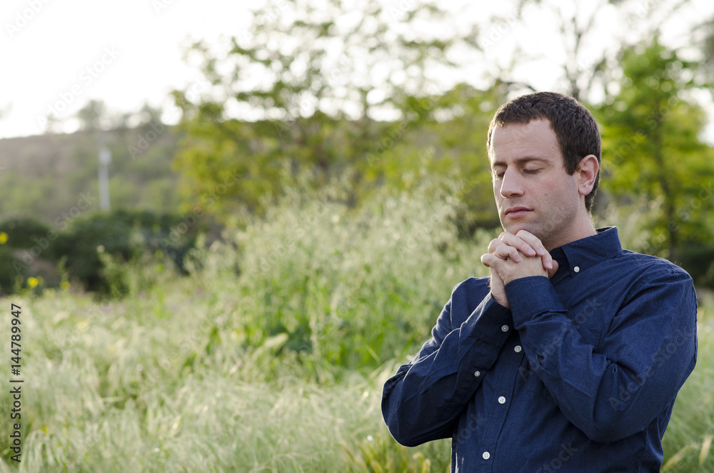 Man praying alone outside in a grassy field with hands folded and eyes closed.