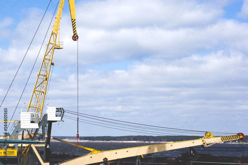 Port crane with yellow arrow against the blue sky and clouds. River port equipment for unloading ships and barges Port crane with yellow arrow against the blue sky and clouds. River port equipment for unloading ships and barges