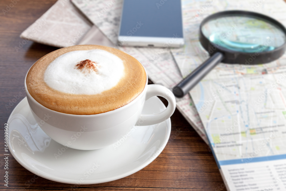 Coffee cup of cappuccino besides with smartphone, maps and magnifier on wooden table