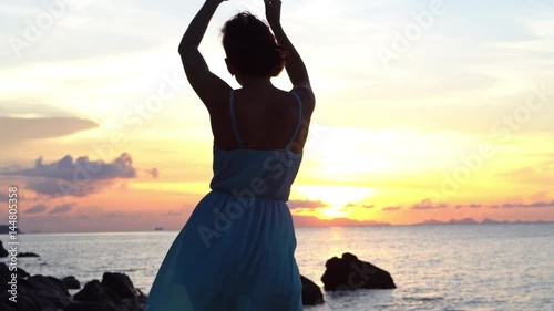 Happy young woman in a long dress dancing on the beach on a colourful sunset background
