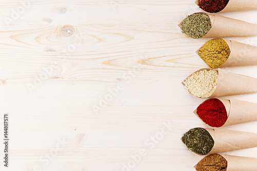 Decorative border of various powder spices close-up in paper corners on white wooden board with copy space.