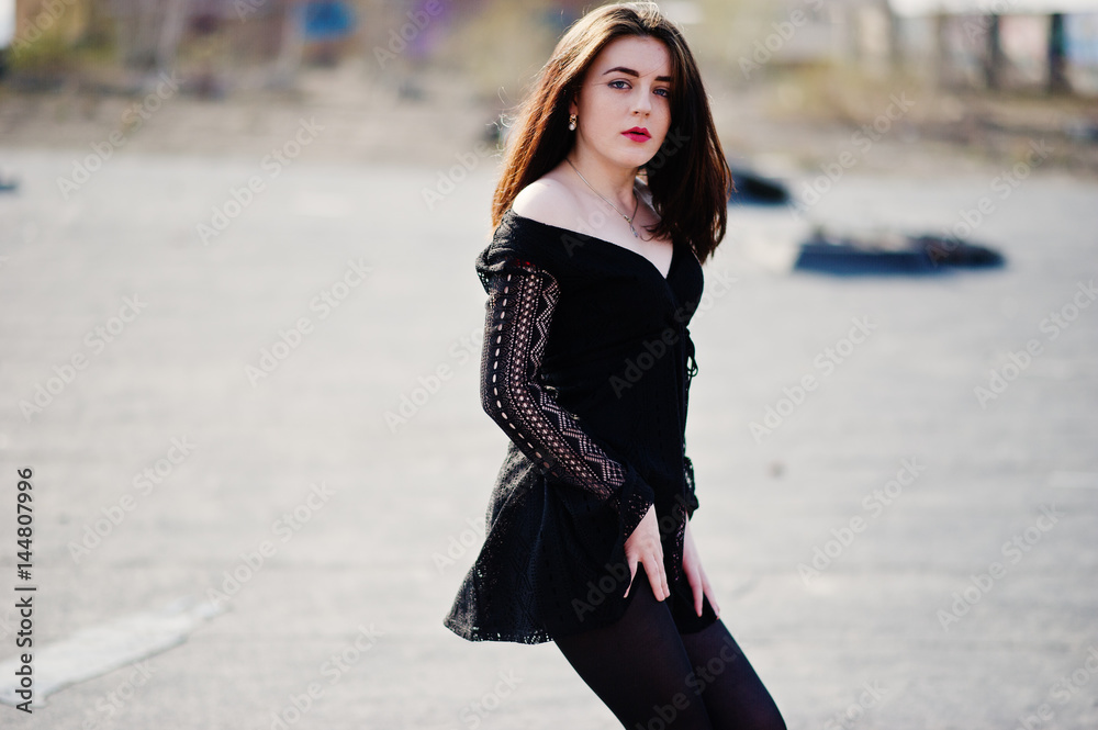 Fototapeta premium Portrait brunette girl with red lips wearing a black dress posed on the roof. Street fashion model.