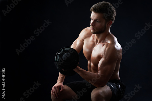 Handsome young sportive shirtless man with muscular strong sexy body doing exercises using dumbbell against black background copyspace gym athletics physique sports motivation achievement gaining. © serhiibobyk