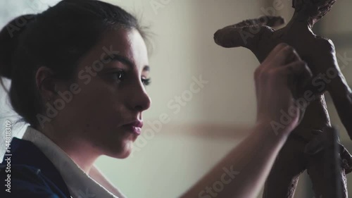 Good young woman makes a sculpture on light art studio with big window. A young student of the art academy makes a clay figure - an ancient statue in the form of a man. Clay figure is an antique man.