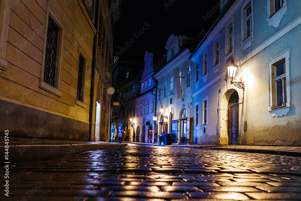 Naklejka premium Oświetlona brukowana ulica z refleksami świetlnymi na chodniku w starym historycznym mieście nocą w Pradze