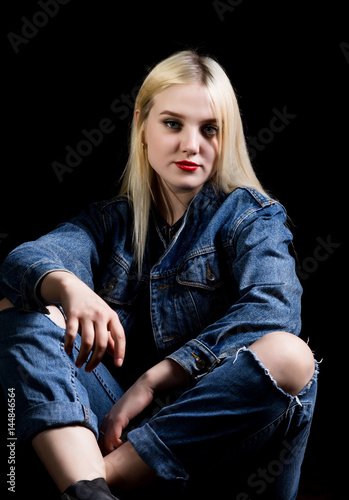 Fashion studio photo of young woman in danim jacket and pants with torn knees sitting on floor, dark background. photo