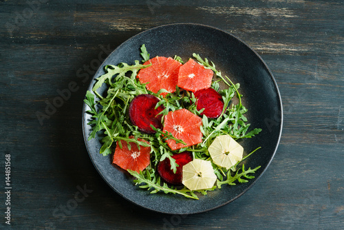 Fresh mix salad of red beet, grapefruit and orange on a cushion of arugula in a black dish on a black wooden table top. View from above.