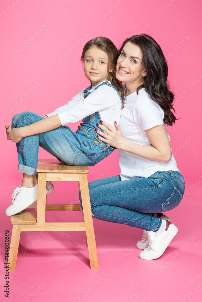 Beautiful happy mother and cute little daughter on stool smiling on pink Beautiful happy mother and cute little daughter on stool smiling on pink