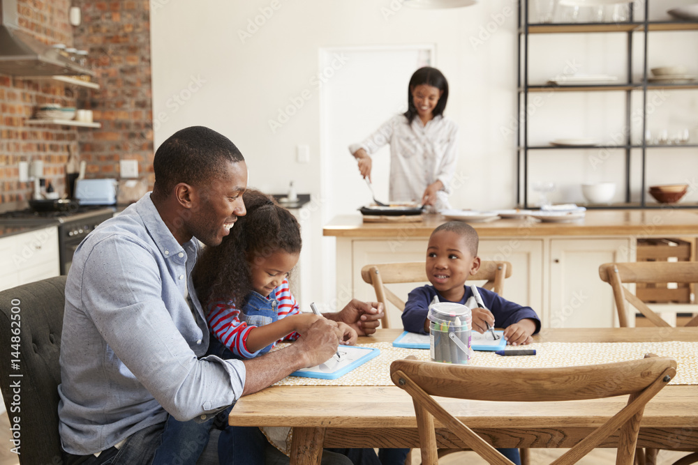Father And Children Drawing At Table As Mother Prepares Meal Father And Children Drawing At Table As Mother Prepares Meal
