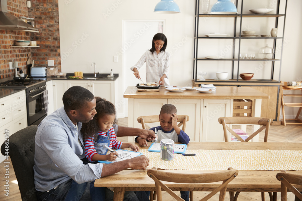 Father And Children Drawing At Table As Mother Prepares Meal