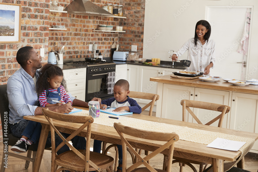 Father And Children Drawing At Table As Mother Prepares Meal Father And Children Drawing At Table As Mother Prepares Meal