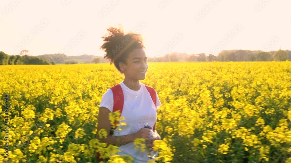 4K video clip of beautiful happy mixed race African American girl teenager female young woman hiking with red backpack and bottle of water in field of rape seed yellow flowers