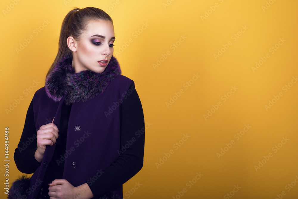 Fototapeta premium Portrait of young gorgeous model with ponytail and artistic make-up wearing trendy purple sleeveless coat with fur collar and looking aside. Isolated on yellow background. Copy-space