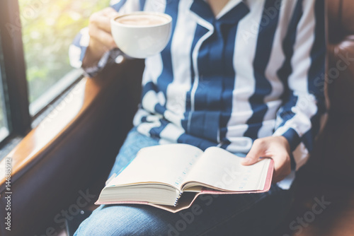 Focus on book. Woman holding a cup of coffee and notebook. Girl reading a book on leather sofa.
