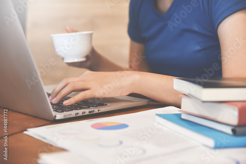 Close up Woman hands holding cup of coffee and using laptop for her work. Graph financial diagram documents on foreground.
