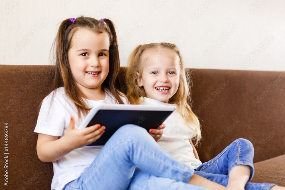Two adorable little sisters playing with a digital tablet at home smiling sister with tablet pc computer Two adorable little sisters playing with a digital tablet at home smiling sister with tablet pc computer
