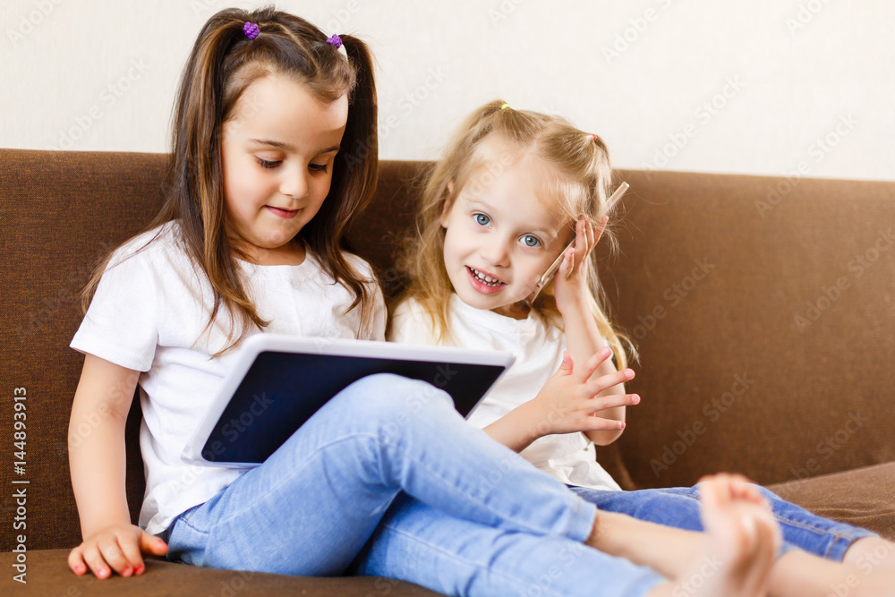 Two adorable little sisters playing with a digital tablet at home smiling sister with tablet pc computer Two adorable little sisters playing with a digital tablet at home smiling sister with tablet pc computer