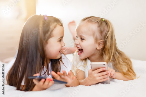 people  children  technology  friends and friendship concept - happy little girls with smartphones lying on floor at home education  school are using a smartphone and smiling while sitting on sofa