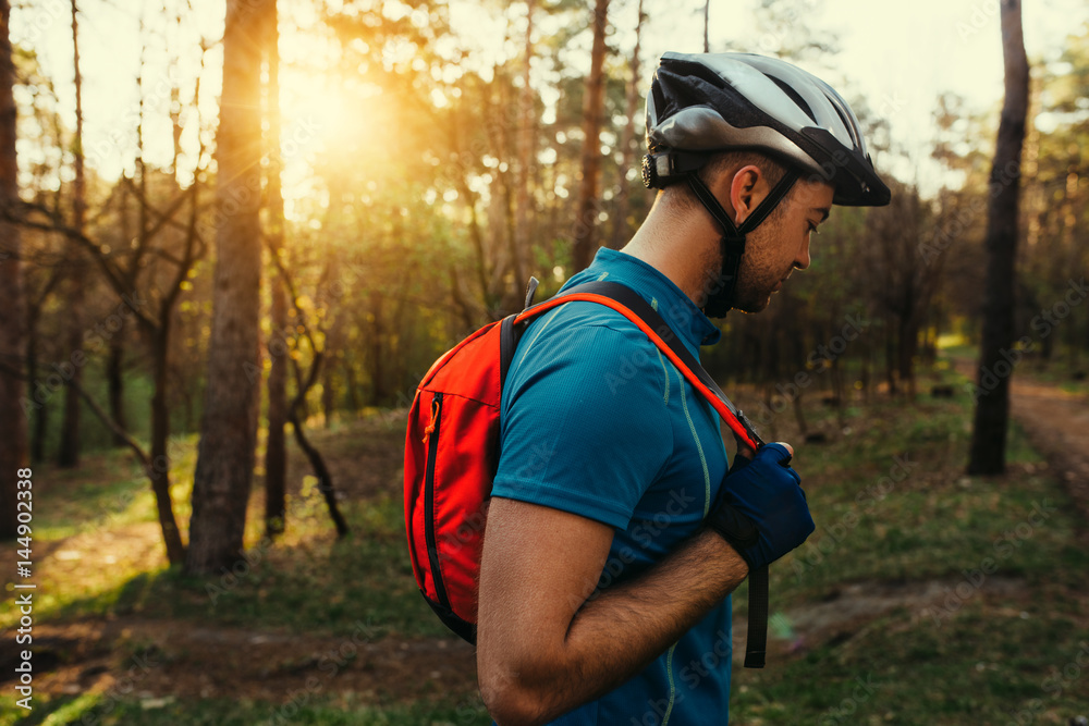 Portrait in profile of handsome young bearded man cyclist wearing protective helmet, blue t-shirt and red backpack, looking down, thinking about route and cycling. Travel concept. Sport lifestyle.