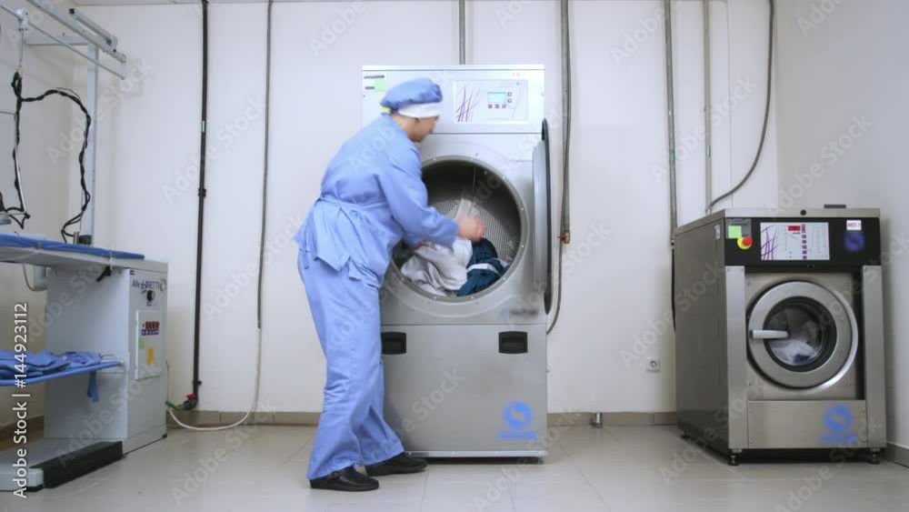 Female worker take away clothes from drying machine at laundry room. Woman removing cloths from washing machine at industrial laundry. Dryer laundry machine in laundry service Female worker take away clothes from drying machine at laundry room. Woman removing cloths from washing machine at industrial laundry. Dryer laundry machine in laundry service