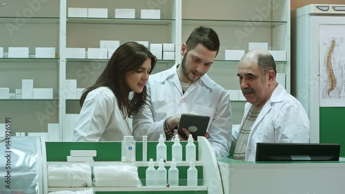 Smiling team of pharmacists, two male and one female, stand side by side in the pharmacy checking information on a tablet computer photo