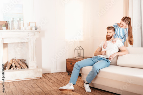 Beautiful young woman hugging handsome bearded man sitting on sofa at home