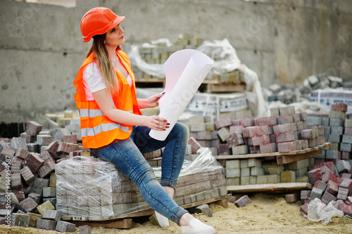 Engineer builder woman in uniform waistcoat and orange protective helmet hold business layout plan paper sitting on pavement.