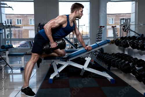 Young muscular man in a T-shirt trains with dumbbells standing with one foot on a bench in the gym