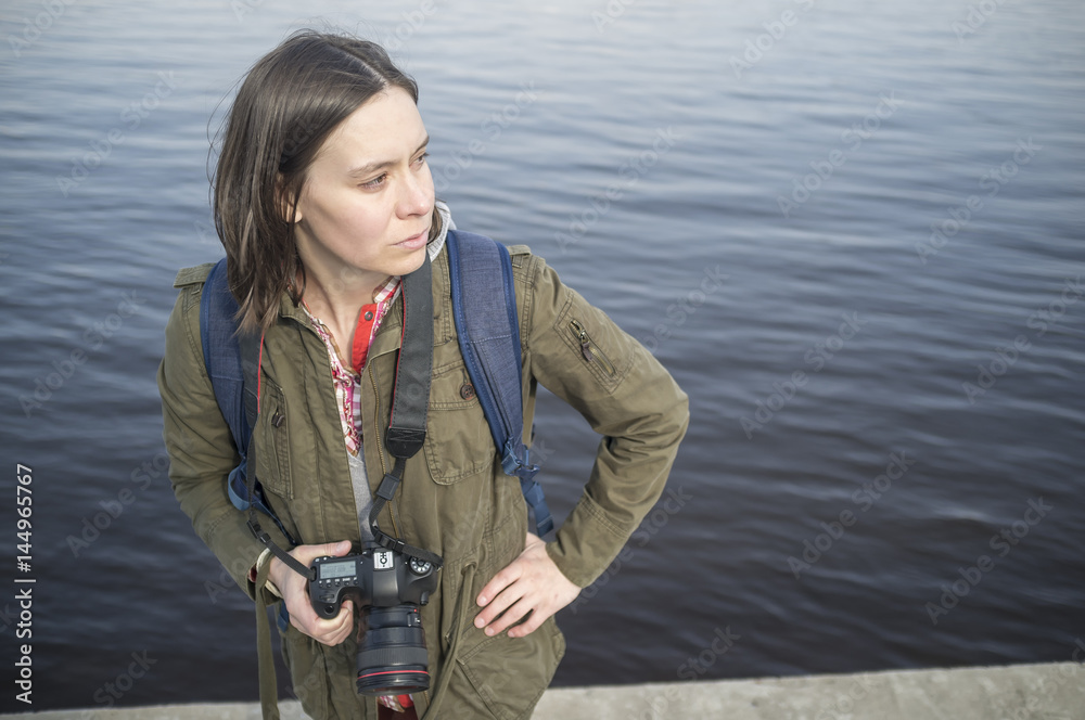 Obraz premium Pensive woman photographer with camera in hand standing on the background of the river and looks into the distance.
