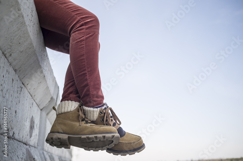 Stylishly dressed girl sits dangling and crossed her legs on a concrete fence against the blue spring sky.