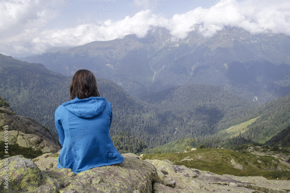 Woman in blue sweater sitting on the edge of a cliff and looking on a beautiful mountain landscape, illuminated by the sun