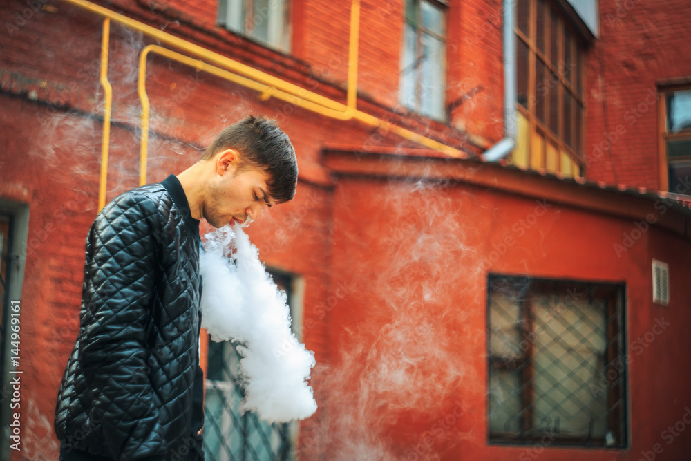 Vape. A handsome young white guy blows steam from an electronic cigarette in a vintage old red yard. Vaping. Vape. A handsome young white guy blows steam from an electronic cigarette in a vintage old red yard. Vaping.