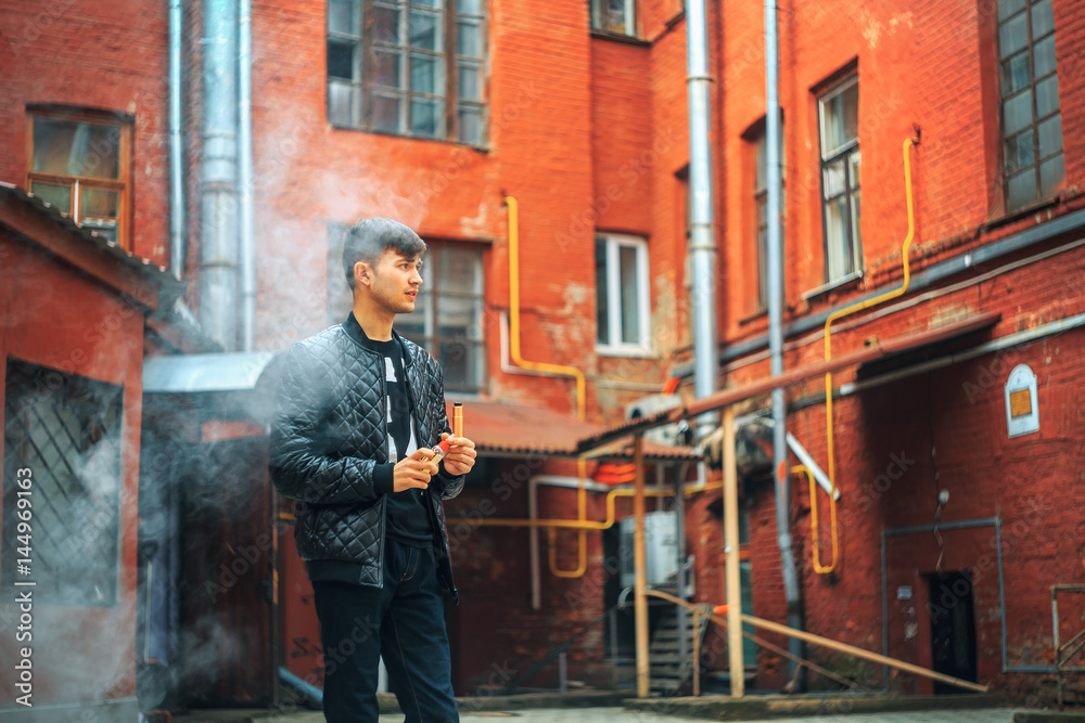 Vape. A handsome young white guy blows steam from an electronic cigarette in a vintage old red yard. Vaping.