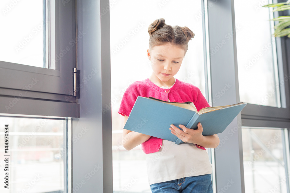 Adorable smiling little girl standing near window and reading book Adorable smiling little girl standing near window and reading book