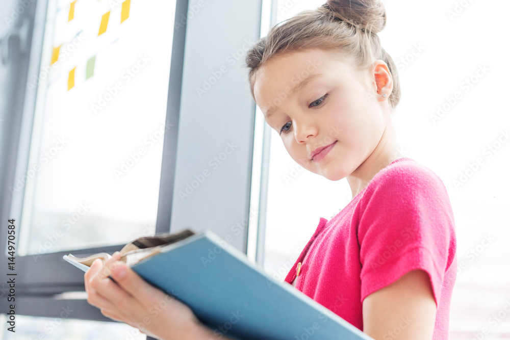 Low angle view of smiling little girl standing near window and reading book Low angle view of smiling little girl standing near window and reading book