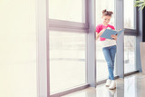 Full length view of smiling little girl standing near window and reading book